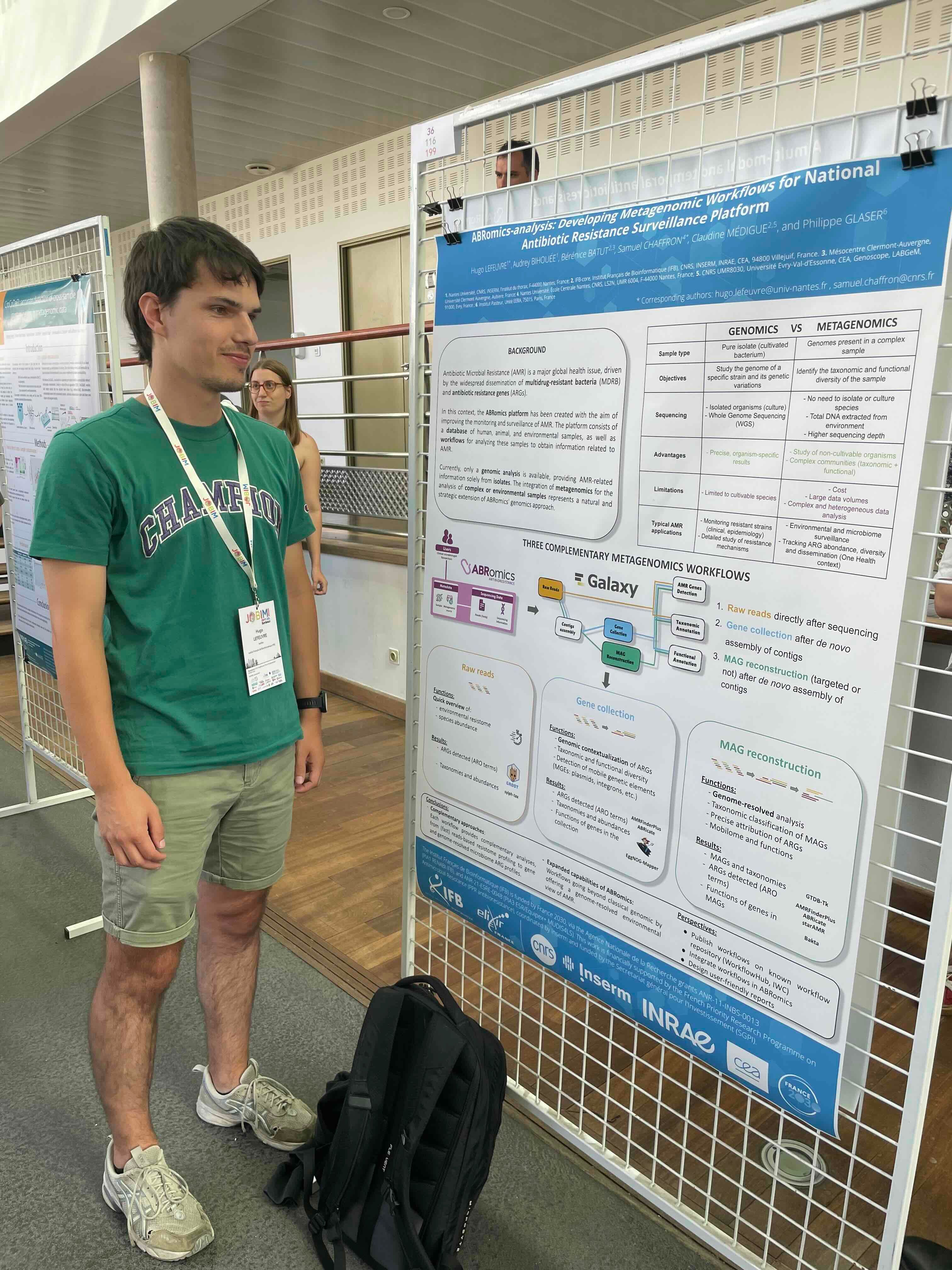The image depicts a scene at a scientific conference. In the foreground, a man wearing a green t-shirt and a conference lanyard stands next to a poster presentation. The poster, titled 'ABRomics-analysis: Developing Metagenomic Workflows for National Antibiotic Resistance Surveillance Platform,' details a study on antibiotic resistance. It compares genomics and metagenomics, outlining their objectives, sequencing methods, advantages, limitations, and applications. The poster also describes three complementary metagenomic workflows: raw reads, gene collection, and MAG (Metagenome-Assembled Genome) reconstruction, all integrated with the Galaxy platform. The background shows a busy conference environment with other attendees and posters, indicating a professional and academic setting focused on scientific research and collaboration.