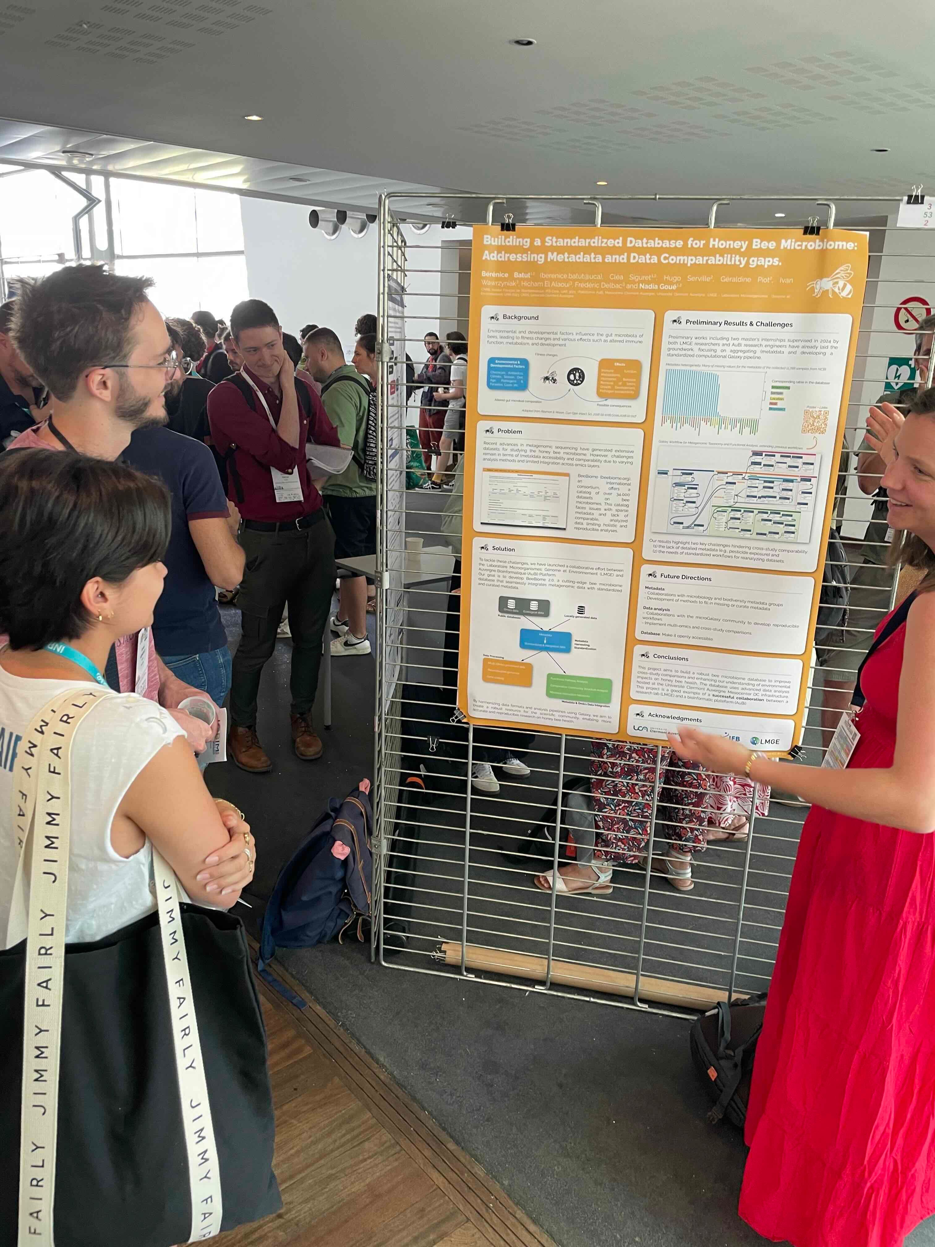 The image depicts a scene at a scientific conference. In the foreground, a group of attendees is gathered around a poster presentation. The poster, titled 'Building a Standardized Database for Honey Bee Microbiome: Addressing Metadata and Data Comparability Gaps,' is displayed on a metal grid. The poster includes sections such as Background, Problem, Solution, Preliminary Results & Challenges, and Future Directions, detailing the research on honey bee microbiomes. The presenters and attendees appear engaged in discussion, with one woman in a red dress pointing to specific parts of the poster, possibly explaining the research findings or methodology. The background shows a busy conference environment with other attendees walking and engaging in discussions, indicating a professional and academic setting focused on scientific research and collaboration.